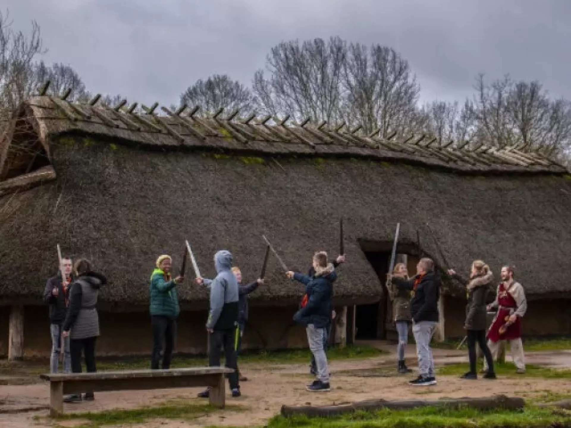 Kinderen oefenen zwaardvechten voor een historisch gebouw met een rieten dak.