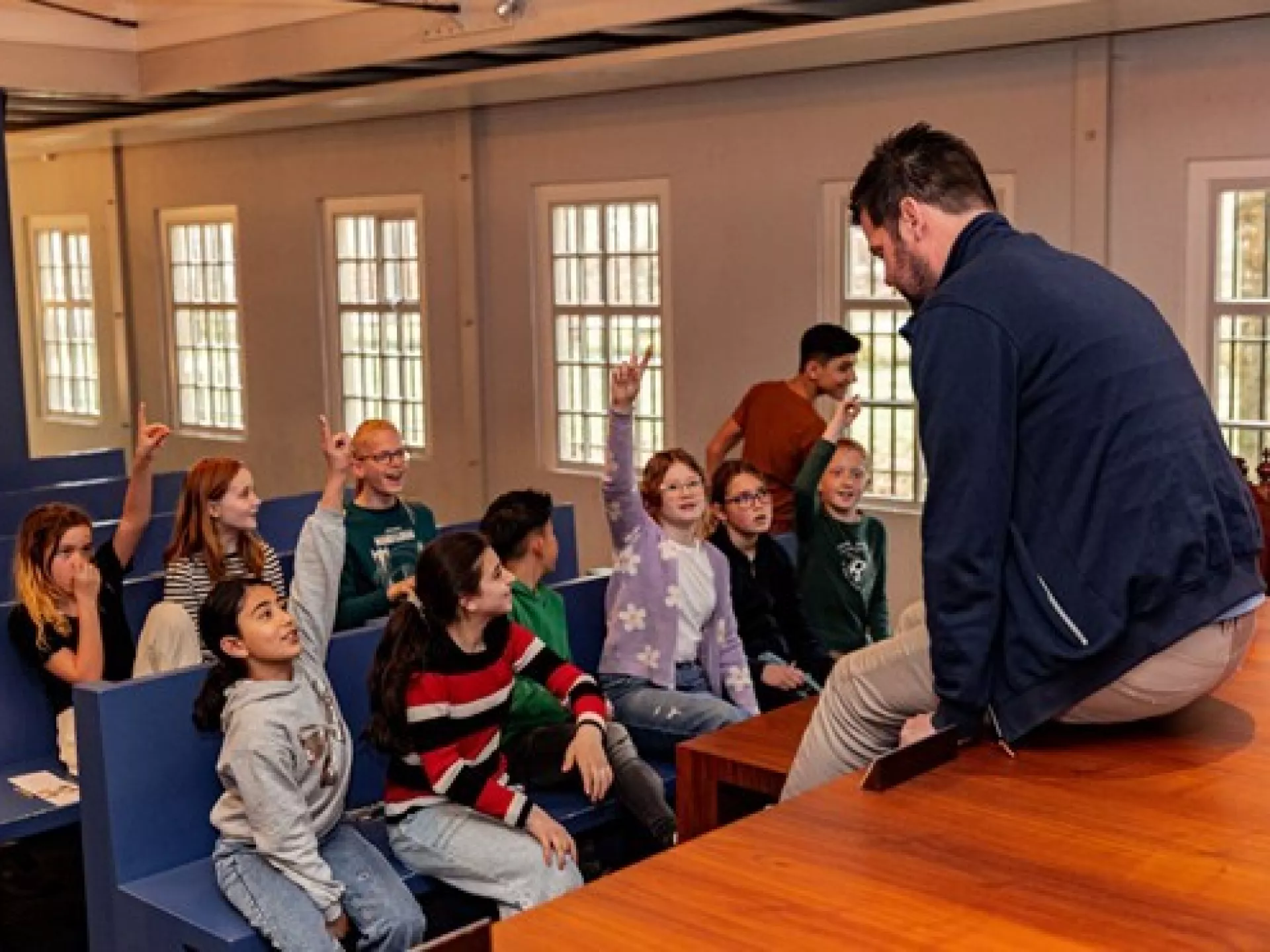 Een groep kinderen luistert naar een spreker die op tafel zit.